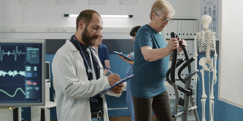 Osteopath taking notes while observing a patient using a stationary bicycle for physical therapy.
