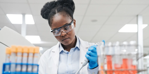 Young female scientist wearing safety goggles and a lab coat, carefully transferring chemical solution using a pipette in a modern laboratory setting.